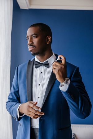 Elegant Black man in blue tuxedo with bow tie applying cologne, standing against blue wall.