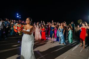 Bride in white lace gown prepares to toss colorful bouquet to excited guests at outdoor evening wedding reception.