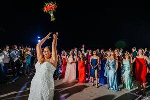 Bride tossing bouquet to excited guests in colorful dresses at outdoor wedding reception at night.