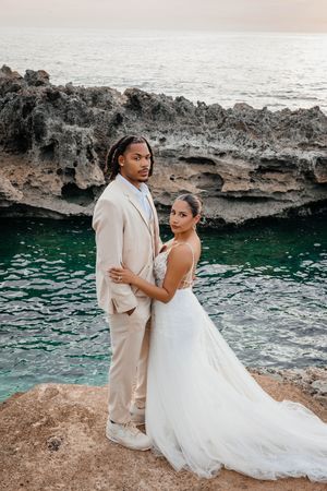 Newlywed couple posing on rocky coastal cliffs with turquoise ocean waters at sunset during wedding photo session.