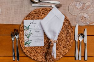 Elegant wedding place setting with woven charger, white napkin, menu card, and silverware on a linen tablecloth.