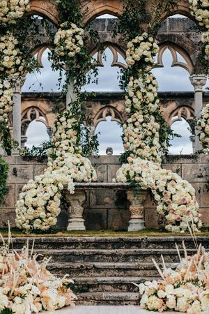 Elegant wedding ceremony altar adorned with lush white floral arches against a Gothic stone venue backdrop.