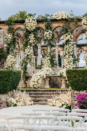 Outdoor wedding ceremony setup featuring white floral arches, stone ruins, and white chairs on a manicured lawn.
