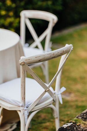 Rustic white cross-back wedding chairs with white cushions and ribbon bows on a green lawn.