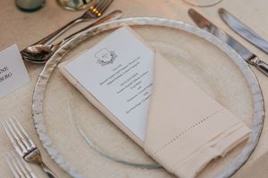 Formal wedding reception place setting with menu card, linen napkin, and silverware on a glass charger plate.