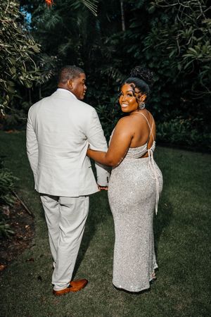 Elegant couple in formal attire posing in a garden, woman in silver sequin gown and man in white suit.