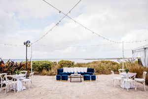 Beachside wedding lounge with navy blue velvet furniture, string lights, and white chairs overlooking ocean views.
