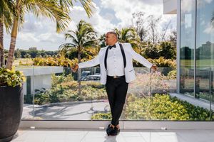 Groom in white tuxedo jacket with black bow tie posing on a terrace surrounded by tropical palms.