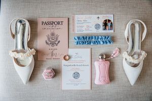 Flat lay of wedding stationery, white heels, pink perfume, blue garter, and ring box on neutral background.