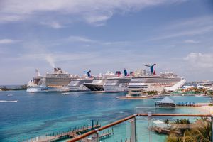 Multiple Carnival cruise ships docked at a tropical port with turquoise waters under a partly cloudy sky.