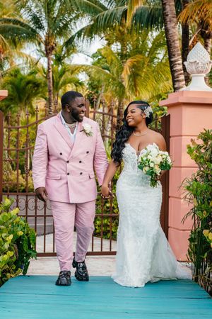 Bride and groom walk hand-in-hand at tropical venue, groom in pink suit, bride in white lace mermaid gown holding bouquet.