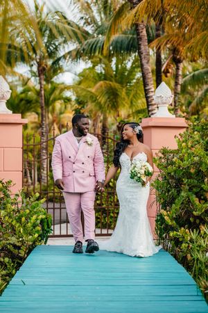 Bride and groom walk hand-in-hand on a turquoise bridge surrounded by palm trees, groom in pink suit, bride in white gown.