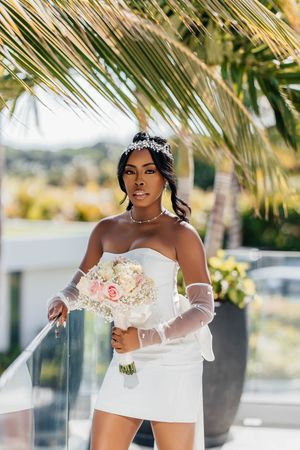 Elegant Black bride in strapless mini dress with tiara holding a blush bouquet under tropical palm fronds.