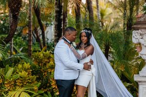 Wedding couple embracing among lush tropical palm gardens, bride wearing white mini dress and cathedral veil with crown.