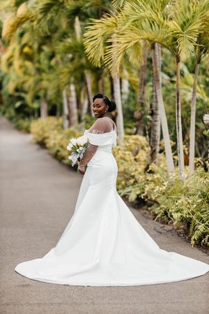 Elegant bride in white mermaid gown with train poses on palm-lined path in tropical garden.