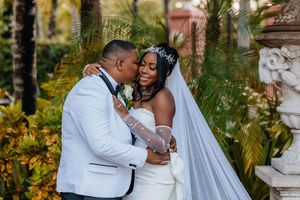 Newlywed couple embracing tenderly among tropical foliage, bride in white gown with veil and headpiece.