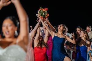 Excited wedding guests reaching up to catch the bridal bouquet toss at an outdoor evening wedding reception.