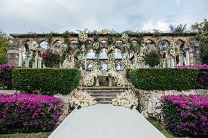 Grand wedding ceremony venue with elaborate white floral arch, stone columns, and vibrant pink bougainvillea hedges.