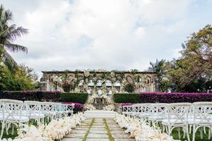 Elegant outdoor wedding ceremony with white chairs, floral aisle, and ivy-covered ruins backdrop.