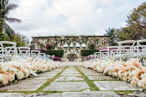 Elegant outdoor wedding ceremony aisle lined with white floral arrangements and chairs at a garden venue.
