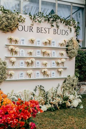 Wedding seating chart display titled Our Best Buds with white flower bundles on a cream wall.