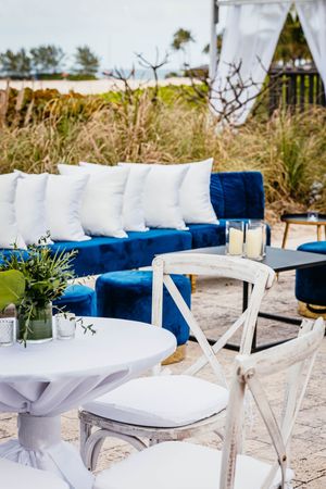Blue velvet outdoor wedding lounge with white pillows, candles, and white bistro tables on a sandy patio.