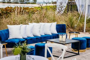 Blue velvet outdoor sofa with white pillows, gold accents, and ottomans surrounding a modern coffee table in a coastal setting.