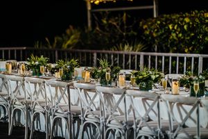 Elegant outdoor wedding reception table with white cross-back chairs, greenery centerpieces, and glowing candles at night.