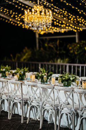 Romantic nighttime outdoor wedding reception with long white tables, greenery centerpieces, and glowing chandelier string lights.
