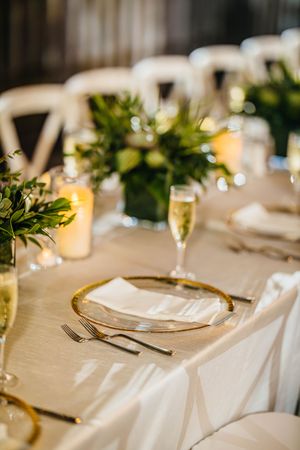 Elegant wedding reception table set with gold-rimmed plates, champagne flutes, candles, and greenery centerpieces.