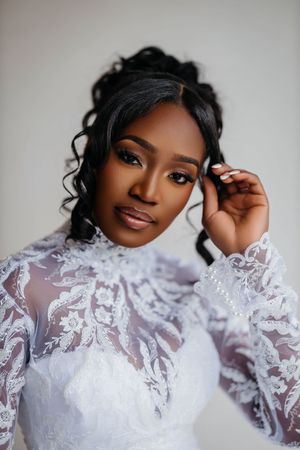 A beautiful Black bride in an elegant white lace high-neck gown with long sleeves poses gracefully against a light background.