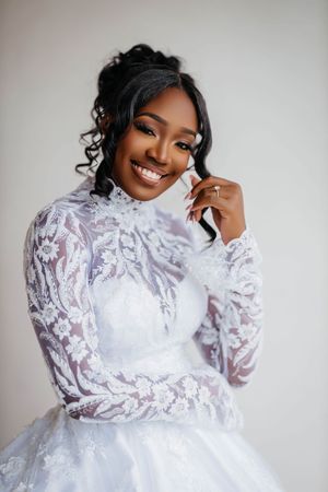 Smiling Black bride in elegant white lace long-sleeve high-neck wedding gown posing gracefully against a light background.