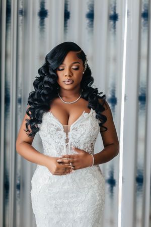 Elegant Black bride in a lace mermaid wedding gown with pearl jewelry and long curly hair, posing gracefully.