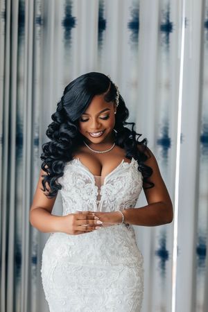 Smiling bride in white lace mermaid gown with diamond necklace and curled hair poses elegantly indoors.