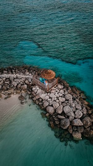 Aerial drone view of a rocky jetty with a thatched umbrella and couple surrounded by turquoise Caribbean waters.