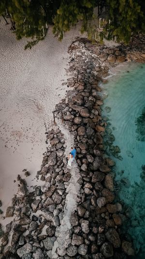 Aerial drone view of a rocky jetty separating turquoise ocean water from sandy beach with two people walking.