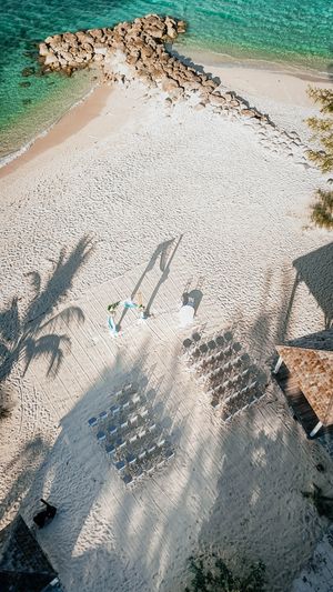Aerial view of a beach wedding ceremony setup with rows of chairs on white sand by turquoise water.