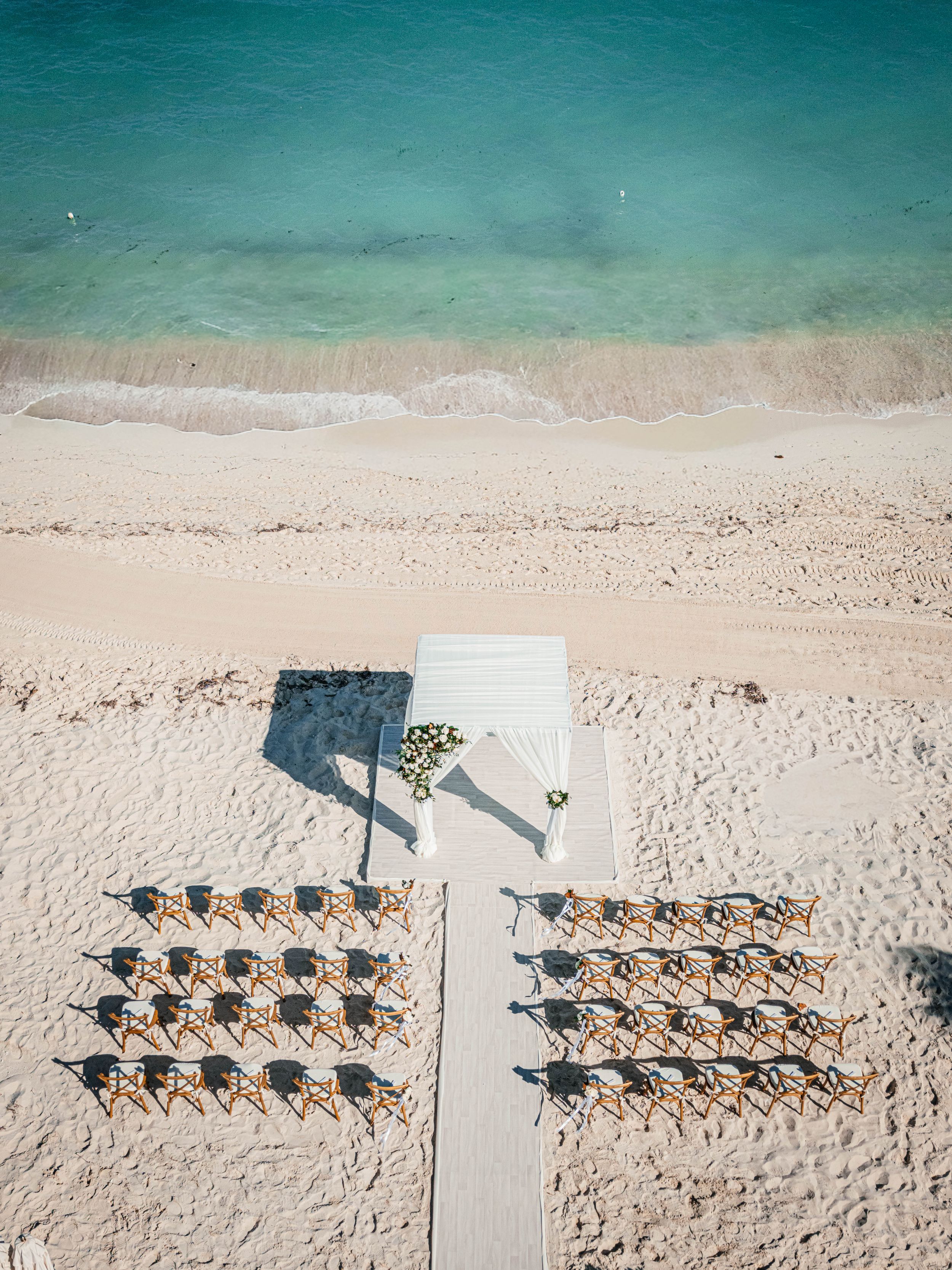 Aerial view of a beach wedding setup with rows of chairs flanking a white aisle leading to an altar by turquoise waters.