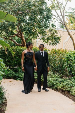 Elegant couple in matching black formal attire walking hand in hand along a garden pathway surrounded by lush greenery.