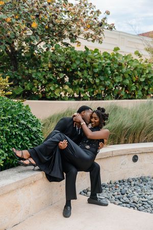 A couple in black formal wear shares a romantic embrace while seated on an outdoor stone wall surrounded by lush greenery.