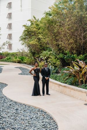 Stylish Black couple in matching black formal attire pose on curved pathway with tropical garden backdrop.
