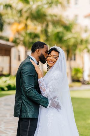 A joyful Black bride in a lace gown and veil embraces her groom in a green suit outdoors.