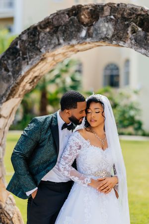 Elegant bride and groom embrace under a stone arch outdoors, bride in lace gown with veil, groom in teal patterned suit.
