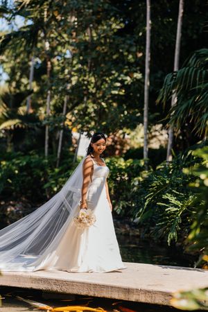 Bride in white gown and veil holds bouquet on dock surrounded by lush tropical greenery near a koi pond.