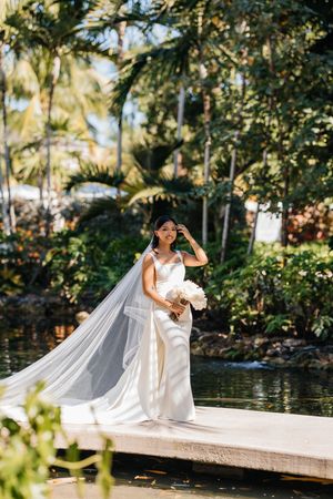 Bride in white gown and long veil holds bouquet on dock surrounded by lush tropical greenery and water.
