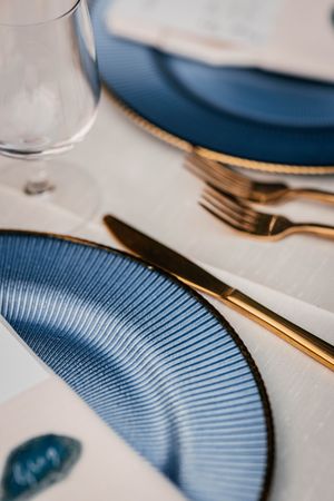 Elegant table setting with blue ribbed plates, gold cutlery, and crystal glassware on a white linen tablecloth.