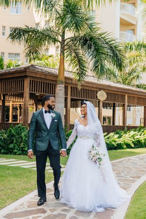 Bride and groom walk hand-in-hand on a tropical resort path, she in lace gown, he in dark tuxedo.