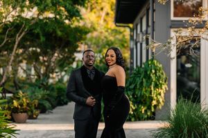 Stylish couple in matching black formal attire smiling together in a lush outdoor garden setting.