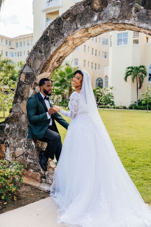 Bride in white lace gown and groom in green suit pose under stone arch at tropical resort wedding.