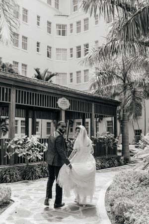Black and white photo of a bride and groom holding hands in a lush courtyard outside an elegant hotel.